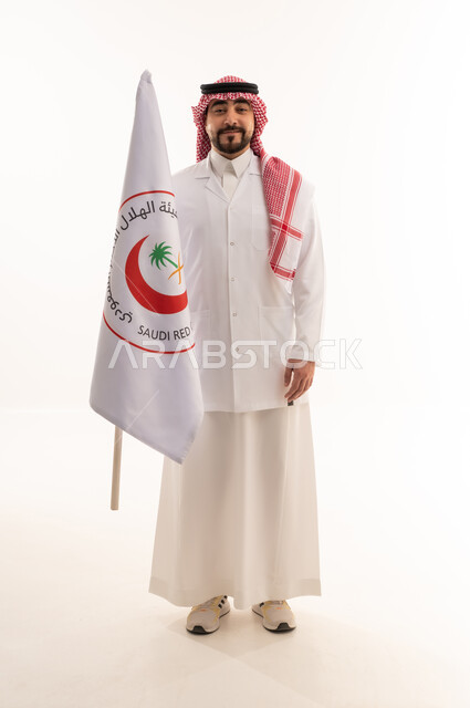 نظرة أمامية للكاميرا مع الابتسامة , طبيب عربي خليجي سعودي يرتدي المعطف الطبي الابيض يحمل راية الهلال الاحمر السعودي , خلفية بيضاء , الجسم بالطول الكامل