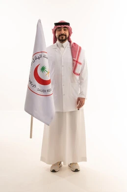 نظرة أمامية للكاميرا مع الابتسامة , طبيب عربي خليجي سعودي يرتدي المعطف الطبي الابيض يحمل راية الهلال الاحمر السعودي , خلفية بيضاء , الجسم بالطول الكامل