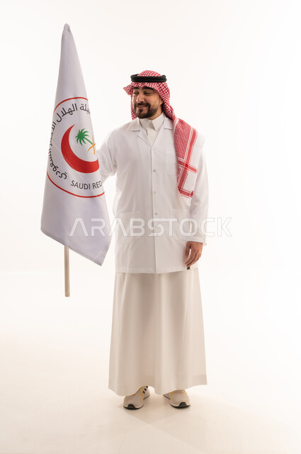 النظر الى علم هيئة الهلال الاحمر السعودي بفخر و اعتزاز , طبيب عربي خليجي سعودي يرتدي المعطف الطبي الابيض يمسك راية الهيئة , خلفية بيضاء , الجسم بالطول الكامل