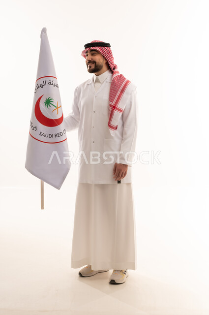 الجسم بالطول الكامل , خلفية بيضاء , نظرة جانبية مع الابتسامة , طبيب عربي خليجي سعودي يرتدي الثوب التقليدي في المملكة مع المعطف الطبي الابيض يرفع علم هيئة الهلال الاحمر السعودي