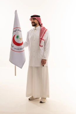 الجسم بالطول الكامل , خلفية بيضاء , نظرة جانبية مع الابتسامة , طبيب عربي خليجي سعودي يرتدي الثوب التقليدي في المملكة مع المعطف الطبي الابيض يرفع علم هيئة الهلال الاحمر السعودي