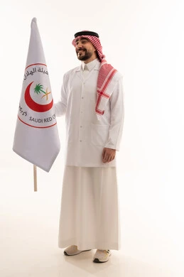 طبيب عربي خليجي سعودي يرتدي المعطف الطبي الابيض يرفع علم هيئة الهلال الاحمر السعودي في سعادة و اعتزاز , خلفية بيضاء , نظرة جانبية , الجسم بالطول الكامل