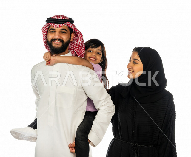 صورة مقربة لرجل عربي خليجي سعودي يقف بجانب زوجته و يحمل ابنته الصغيرة على ظهره بإيماءات تدل على السعادة و المحبة ، أجواء عائلية سعيدة ، مفهوم الأسرة السعيدة ، قضاء أوقات عائلية ممتعة