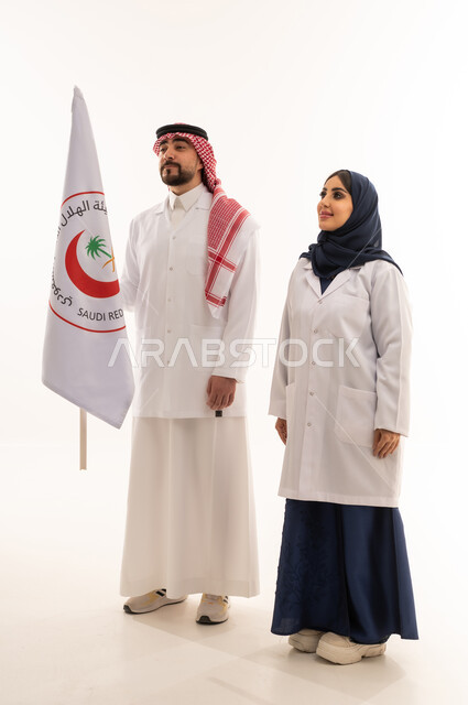 صورة تذكارية لاطباء هيئة الهلال الاحمر السعودي , طبيبة عربية خليجية سعودية محجبة ترتدي عباءة زرقاء و معطف طبي ابيض تقف بجانب زميل العمل , طبيب عربي خليجي سعودي يرتدي المعطف الطبي الابيض يرفع علم هيئة الهلال الاحمر السعودي بفخر و اعتزاز , خلفية بيضاء, الجسم بالطول الكامل , النظر الى شئ ما