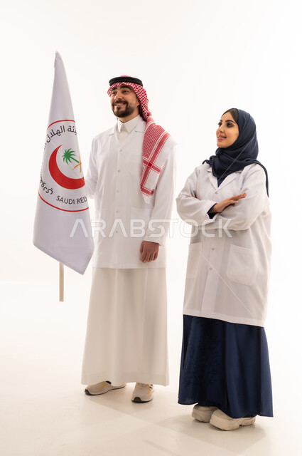 طبيب عربي خليجي سعودي يرتدي المعطف الطبي الابيض و الثوب التقليدي للمملكة يرفع علم هيئة الهلال الاحمر السعودي في عزة و فخر , طبيبة عربية خليجية سعودية محجبة ترتدي عباءة زرقاء و معطف طبي ابيض تقف عاقدة ذراعيها بجانب الطبيب , النظر الى شئ ما , خلفية بيضاء, الجسم بالطول الكامل