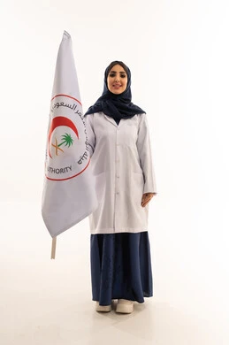 A front-facing look at the camera with a smile, an Arab Gulf Saudi female doctor wearing a white medical coat standing next to the flag of the Saudi Red Crescent Authority, a symbol of distinguished competencies, portrait with a white background, full-body.