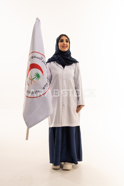 طبيبة عربية خليجية سعودية محجبة ترتدي عباءة زرقاء و المعطف الطبي الابيض ترفع علم هيئة الهلال الاحمر السعودي بسعادة بالغة , خلفية بيضاء , الجسم بالطول الكامل , النظر للاعلى