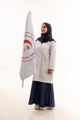 ايماءات السعادة واضحة على وجه الطبيبة , طبيبة عربية خليجية محجبة ترتدي عباءة زرقاء اللون تقف بجانب علم هيئة الهلال الاحمر السعودي بفخر , نظرة جانبية ,خلفية بيضاء, الجسم بالطول الكامل