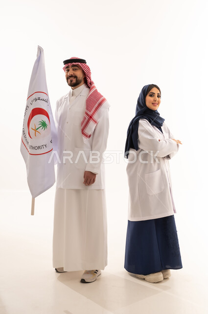 Doctors of the Saudi Red Crescent Authority, an Arab Gulf Saudi doctor wearing a white medical coat and the traditional dress of the Kingdom stands opposite the female doctor, an Arab Gulf Saudi female doctor in a hijab wearing a blue abaya stands proudly next to the Authority's flag, looking directly at the camera, full body shot, white background.