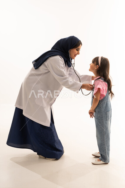 تبادل الابتسامات و الضحك بين الطبيبة و الفتاة الصغيرة , طفلةعربية خليجية سعودية تقف امام الطبيبة مبتسمة اثناء عملية الفحص الطبي , طبيبة عربية خليجية محجبة ترتدي المعطف الطبي الابيض تستمع الى دقات قلب الصغيرة , بورتريه خلفية بيضاء , الجسم بالطول الكامل
