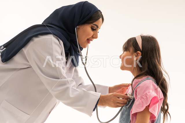 طبيبة القلب العربية الخليجية السعودية تنحني امام الفتاة الصغيرة لاجراء الفحص الدوري اللازم , طفلة عربية خليجية سعودية ترتدي الزي الكاجوال تقف امام الطبيبة مبتسمة اثناء اجراء الكشف , بورتريه مقرب , خلفية بيضاء