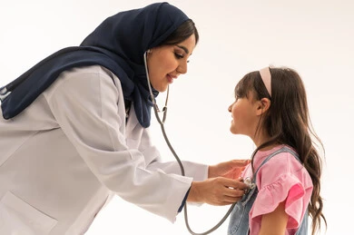 طبيبة القلب العربية الخليجية السعودية تنحني امام الفتاة الصغيرة لاجراء الفحص الدوري اللازم , طفلة عربية خليجية سعودية ترتدي الزي الكاجوال تقف امام الطبيبة مبتسمة اثناء اجراء الكشف , بورتريه مقرب , خلفية بيضاء