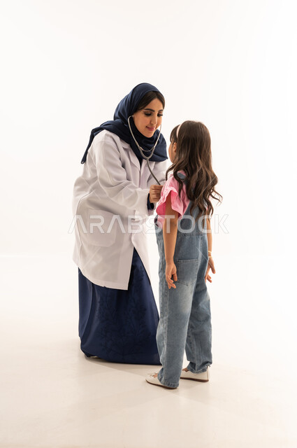 ممارسة مهنة الطب بحرفية تامة , طبيبة قلب عربية خليجية سعودية ترتدي المعطف الطبي الابيض تجري فحصاً دورياً للفتاة الصغيرة , طفلة عربية خليجية سعودية ترتدي الزي الكاجوال تقف امام الطبيبة لاستكمال عملية الفحص , بورتريه خلفية بيضاء, الجسم بالطول الكامل