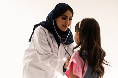 An Arab Gulf Saudi female doctor wearing a blue abaya and a white medical coat is using a stethoscope to listen to the heartbeats of patients. An Arab Gulf Saudi girl dressed in casual attire stands in front of the doctor for important examinations. Close-up portrait, white background.