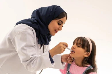 طبيبة عربية خليجية سعودية محجبة ترتدي عباءة زرقاء و معطف طبي ابيض تنحي امام الفتاة الصغيرة لمتابعة حالتها الصحية ,طفلة عربية خليجية سعودية ترتدى الزي الكاجوال تقيس درجة حرارة جسدها ,بورتريه مقرب , خلفية بيضاء