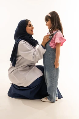 Performing medical tasks with professionalism and kindness, an Arab Gulf Saudi female doctor wearing a white medical coat squats in front of a small girl to examine her heart. The Arab Gulf Saudi girl, dressed in casual attire, stands in front of the doctor to complete the examination process. Portrait with a white background, full-body shot.