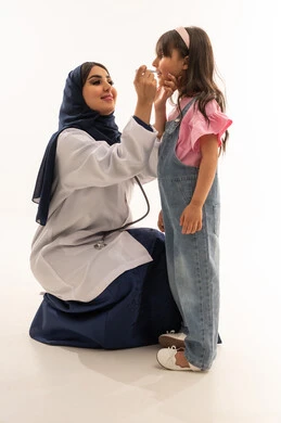 Monitoring the health status of children and all patients of all ages, an Arab Gulf Saudi female doctor wearing a white medical coat squats in front of a little girl, an Arab Gulf Saudi child wearing casual attire measuring body temperature, portrait with a white background, full body.