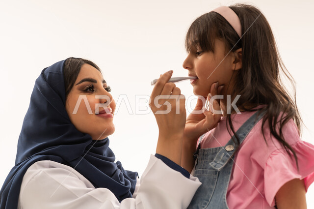Monitoring the normal body temperature is one of the essential necessities during children's examinations. An Arab Gulf Saudi female doctor wearing a white medical coat holds a thermometer, while an Arab Gulf Saudi girl dressed in casual attire stands firmly in front of the doctor. Close-up portrait, white background.