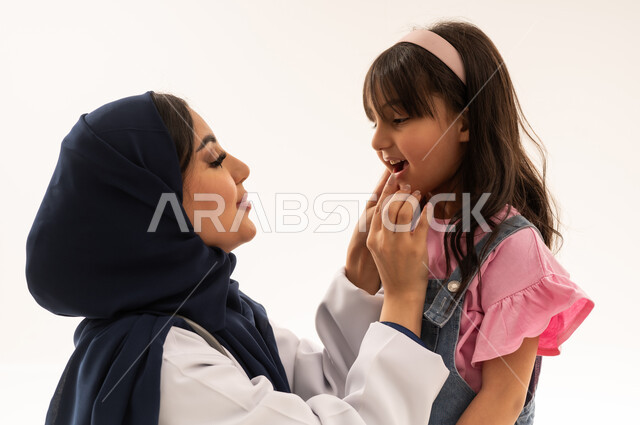 طبيبة عربية خليجية سعودية محجبة ترتدي المعطف الطبي الابيض تجلس القرفصاء امام الفتاة الصغيرة , فحص دوري على الاسنان و اللثة , طفلة عربية خليجية سعودية ترتدي الزي الكاجوال تقف امام الطبيبة للقيام بفحص الاسنان , بورتريه مقرب , خلفية بيضاء