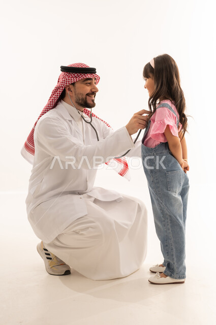 التعامل بلطف و محبة مع المرضى , طبيب عربي خليجي سعودي يرتدي المعطف الابيض الطبي يجلس القرفصاء امام الفتاة الصغيرة , طفلة عربية خليجية سعودية ترتدي الزي الكاجوال تقوم بالفحص الطبي للقلب , بورتريه خلفية بيضاء , الجسم بالطول الكامل