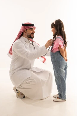Dealing with kindness and love with patients, an Arab Gulf Saudi doctor wearing a white medical coat squats in front of a little girl, an Arab Gulf Saudi child wearing casual attire conducting a heart examination, portrait with a white background, full body.