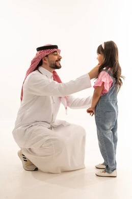 An Arab Gulf Saudi doctor wearing a white medical coat sits in front of a squatting little girl, gestures of happiness and smiling. An Arab Gulf Saudi girl in casual attire stands in front of the dentist, happy after completing her treatment, with a white background, full-body portrait.