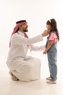 طبيب عربي خليجي سعودي يرتدي المعطف الطبي الابيض يجلس القرفصاء امام الفتاة الصغيرة في مداعبة لطيفة , طبيبة عربية خليجية سعودية ترتدي الزي الكاجوال تبتسم للطبيب الكف بعد الـاطمئنان على الصحة . بورتريه خلفية بيضاء , الجسم بالطول الكامل