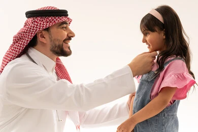 A close-up portrait filled with joy and happiness between the doctor and the little girl. An Arab Gulf Saudi doctor wearing a white medical coat is joking with the girl. The Arab Gulf Saudi girl is wearing casual attire and smiling at the doctor after the examination and reassurance about her general health, with a white background.