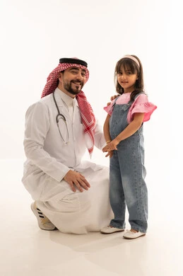 صورة تذكارية تكشف علاقة الود و المحبة بين الطبيب و المرضى , طبيب عربي خليجي سعودي يرتدي المعطف الطبي الابيض يجلس قرفصاء بجانب فتاة صغيرة , طفلة عربية خليجية سعودية ترتدي الزي الكاجوال تقف بجانب الطبيب بعد اتمام الفحص و العلاج , بورتريه خلفية بيضاء , الجسم بالطول الكامل