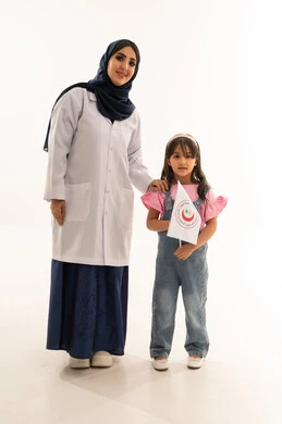 طفلة عربية خليجية سعودية ترتدي الزي الكاجوال ترفع علم الهلال الاحمر السعودي , طبيبة عربية خليجية سعودية محجبة ترتدي المعطف الطبي الابيض تقف بجانب الفتاة الصغيرة , النظر للكاميرا , بورتريه خلفية بيضاء , الجسم بالطول الكامل