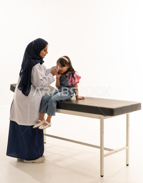 التعامل مع الحالات الحرجة بحرفية تامة ,طبيبة عربية خليجية سعودية ترتدي المعطف الطبي الابيض تقوم بنزع الضمادة بهدوء لتجنب الاحساس بالالم , طفلة عربية خليجية سعودية ترتدي الزي الكاجوال تجلس على سرير الكشف واثقة في طبيبتها , بورتريه خلفية بيضاء , الجسم بالطول الكامل