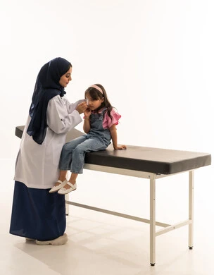 Dealing with critical cases with complete professionalism, an Arab Gulf Saudi doctor wearing a white medical coat gently removes the bandage to avoid causing pain. An Arab Gulf Saudi girl dressed in casual attire sits confidently on the examination bed, trusting her doctor. Portrait with a white background, full-body.