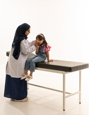 Post-surgical healthcare is required. A Saudi Arabian Gulf Arab female doctor wearing a white medical coat is removing the medical bandage from the girl's eye. A Saudi Arabian Gulf Arab girl dressed in casual attire sits steadily and confidently during the follow-up after the surgery. Portrait with a white background, full-body shot.