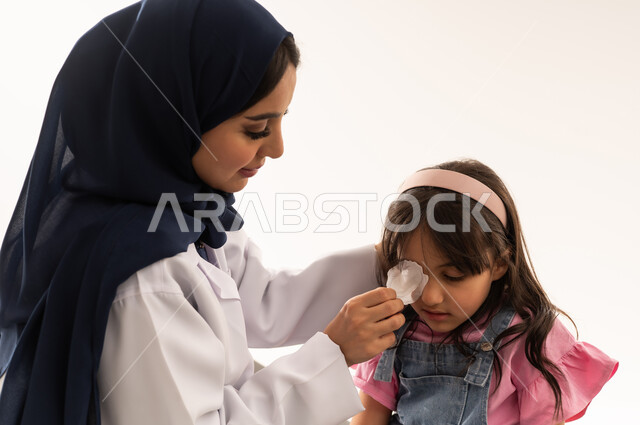 كفاءات هيئة الهلال الاحمر السعودي تقوم باجراء العمليات الجراحية الحرجة باحترافية , طبيبة عربية خليجية سعودية محجبة ترتدي معطف طبي ابيض تنزع الضمادة من عين الفتاة الصغيرة , طفلة عربية خليجية سعودية ترتدي الزي الكاجوال تجلس على سرير الكشف بعد اجراء عملية حرجة , بورتريه مقرب , خلفية بيضاء
