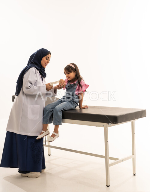 ايماءات السعادة اثناء تلقي العلاج المناسب , طفلة عربية سعودية خليجية تجلس على سرير الكشف لوضع الضمادة على ذراعها , طبيبة عربية خليجية سعودية محجبة ترتدي المعطف الطبي الابيض تقوم بممارسة عملها بكفاءة , بورتريه خلفية بيضاء , الجسم بالطول الكامل
