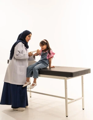 ايماءات السعادة اثناء تلقي العلاج المناسب , طفلة عربية سعودية خليجية تجلس على سرير الكشف لوضع الضمادة على ذراعها , طبيبة عربية خليجية سعودية محجبة ترتدي المعطف الطبي الابيض تقوم بممارسة عملها بكفاءة , بورتريه خلفية بيضاء , الجسم بالطول الكامل