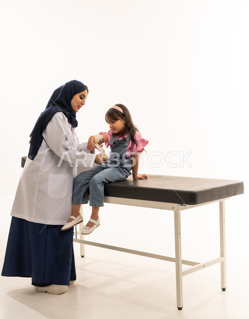 جميع التخصصات متوافرة بكفاءة في هيئة الهلال الاحمر السعودي , طبيبة عربية خليجية سعودية محجبة ترتدي معطف ابيض طبي تقوم بتقديم العلاج المناسب للفتاة الصغيرة , طفلة عربية خليجية سعودية تجلس على سرير الكشف اثناء وضع الضمادة على ذراعها , بورتريه خلفية بيضاء , الجسم بالطول الكامل