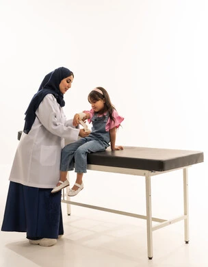 جميع التخصصات متوافرة بكفاءة في هيئة الهلال الاحمر السعودي , طبيبة عربية خليجية سعودية محجبة ترتدي معطف ابيض طبي تقوم بتقديم العلاج المناسب للفتاة الصغيرة , طفلة عربية خليجية سعودية تجلس على سرير الكشف اثناء وضع الضمادة على ذراعها , بورتريه خلفية بيضاء , الجسم بالطول الكامل