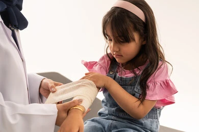 ايماءات حزينة خائفة على وجه الفتاة الصغيرة , طفلة عربية خليجية سعودية تشعر بالالم اثناء وضع الضمادة , طبيبة عربية خليجية سعودية محجبة ترتدي معطف طبي ابيض تقوم بمعالجة المريض بالطريقة المناسبة لها , بورتريه مقرب , خلفية بيضاء