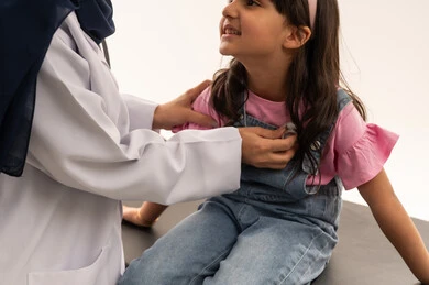 السعادة و الابتسامة و الثقة المتبادلة بين الاطباء و المريض , طبيبة عربية خليجية سعودية محجبة ترتدي المعطف الطبي الابيض تفحص قلب الفتاة الصفيرة , طفلة عربية خليجية سعودية ترتدي الزي الكاجوال تداعب الطبيبة اثناء الفحص الدوري , خلفية بيضاء , بورتريه مقرب