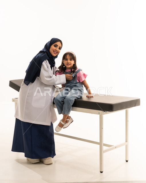 النظر للكاميرا مع ايماءات السعادة و الطمأنينة ,طبيبة عربية خليجية سعودية محجبة ترتدي المعطف الابيض الطبي تستمع بانصات الى نبضات قلب الفتاة الصغيرة , طفلة عربية خليجية سعودية ترتدي الزي الكاجوال تجلس على سرير الكشف اثناء الفحص الدوري للقلب , بورتريه خلفية بيضاء , الجسم بالطول الكامل