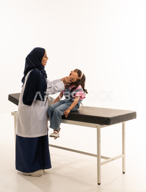 الحفاظ على الاسنان من التسوس منذ الصغر  , طفلة عربية خليجية سعودية ترتدي الزي الكاجوال تجلس في عيادة الاسنان للقيام بالفحص الدوري للفم و اللثة , طبيبة اسنان عربية خليجية سعودية محجبة ترتدي المعطف الابيض الطبي تقوم بفحص اسنان الفتاة الصغيرة , بورتريه خلفية بيضاء , الجسم بالطول الكامل