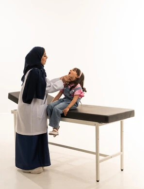الحفاظ على الاسنان من التسوس منذ الصغر  , طفلة عربية خليجية سعودية ترتدي الزي الكاجوال تجلس في عيادة الاسنان للقيام بالفحص الدوري للفم و اللثة , طبيبة اسنان عربية خليجية سعودية محجبة ترتدي المعطف الابيض الطبي تقوم بفحص اسنان الفتاة الصغيرة , بورتريه خلفية بيضاء , الجسم بالطول الكامل