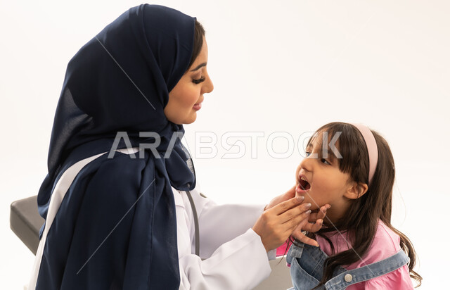 العناية بالاسنان منذ الصغر والحفاظ عليها من التسوس , طفلة عربية خليجية سعودية ترتدي الزي الكاجوال تجلس على سرير الكشف في عيادة فحص الاسنان , طبيبة عربية خليجية سعودية محجبة ترتدي المعطف الطبي الابيض تقوم بالكشف عن امراض اللثة و الفم ,بورتريه مقرب , خلفية بيضاء
