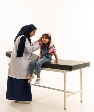 التركيز اثناء القيام بفحص الاذن و حاسة السمع , طفلة عربية خليجية سعودية  ترتدي الزي الكاجوال تجلس على سرير الكشف لتشخيص الحالة , طبيبة عربية خليجية سعودية محجبة ترتدي المعطف الطبي الابيض تقوم بعملها باحترافية تامة , بورتريه خلفية بيضاء , الجسم بالطول الكامل