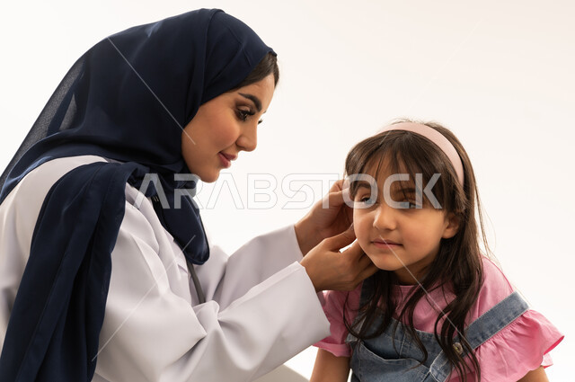 الكشف المبكرع على الاطفال لاكتشاف الامراض و ضعف حاسة السمعمن المهام الرئيسية المتاحة في هيئة الهلال الاحمر السعودي , طبيبة عربية خليجية سعودية محجبة ترتدي المعطف الطبي الابيض تفحص أذن الفتاة الصغيرة , طفلة عربية خليجية سعودية ترتدي الزي الكاجوال تجلس على سرير الكشف لتلقي المساعدة اللازمة , بورتريه مقرب ,خلفية بيضاء