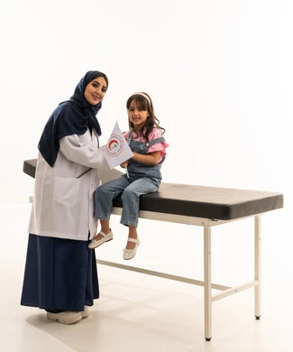 The assistance of experienced and qualified doctors at the Saudi Red Crescent Authority, an Arab Gulf Saudi female doctor in a hijab wearing a white medical coat holding the organization's flag, a small Arab Saudi girl sitting on the examination bed, portrait with a white background, full body.