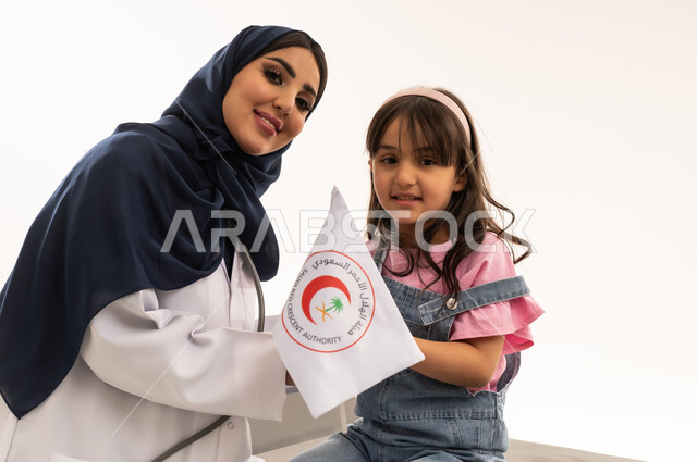 علاقات المودة والثقة في اطباء هيئة الهلال الاحمر السعودي, طبيبة عربية خليجية سعودية محجبة ترتدي المعطف الطبي الابيض تقف بجانب الفتاة الصغيرة ,طفلة عربية خليجية سعودية ترتدي الزي الكاجوال تحمل راية الهلال الاحمر السعودي بفخر و محبة , بورتريه مقرب,خلفية بيضاء , النظر للكاميرا