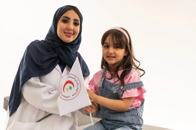 The flag of the Saudi Red Crescent Authority is raised as a symbol of trust and efficiency. A veiled Arab Gulf Saudi female doctor wearing a white medical coat stands next to a small girl dressed in casual attire, both happily holding the flag. The background is white, and it is a close-up portrait.