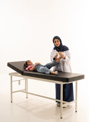 ممارسة مهنة الطب باحترافية , طبيبة عربية خليجية سعودية محجبة ترتدي المعطف الطبي الابيض تقدم العلاج اللازم للفتاة الصغيرة , طفلة عربية خليجية سعودية نائمة على سرير الكشف ترفع قدمها المصابة لتلقي المساعدة , بورتريه خلفية بيضاء , الجسم بالطول الكامل